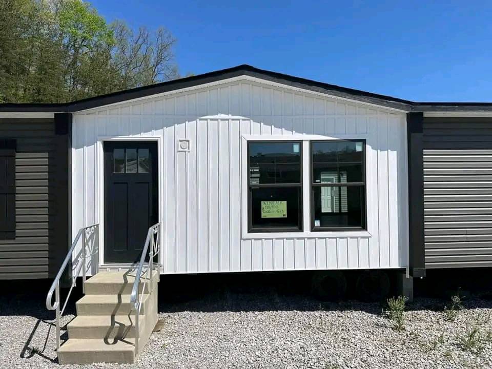 A modern one-story modular home with a white facade and gray sides. It has a glass-panel front door, three-step entry with metal railings, and a large window, set against a backdrop of clear blue sky and trees.