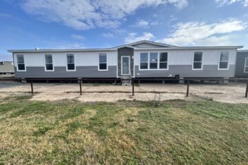 A single-story manufactured home with a gray and white exterior, featuring large windows and a central door. It sits on a gravel area with grass in the foreground under a partly cloudy blue sky.
