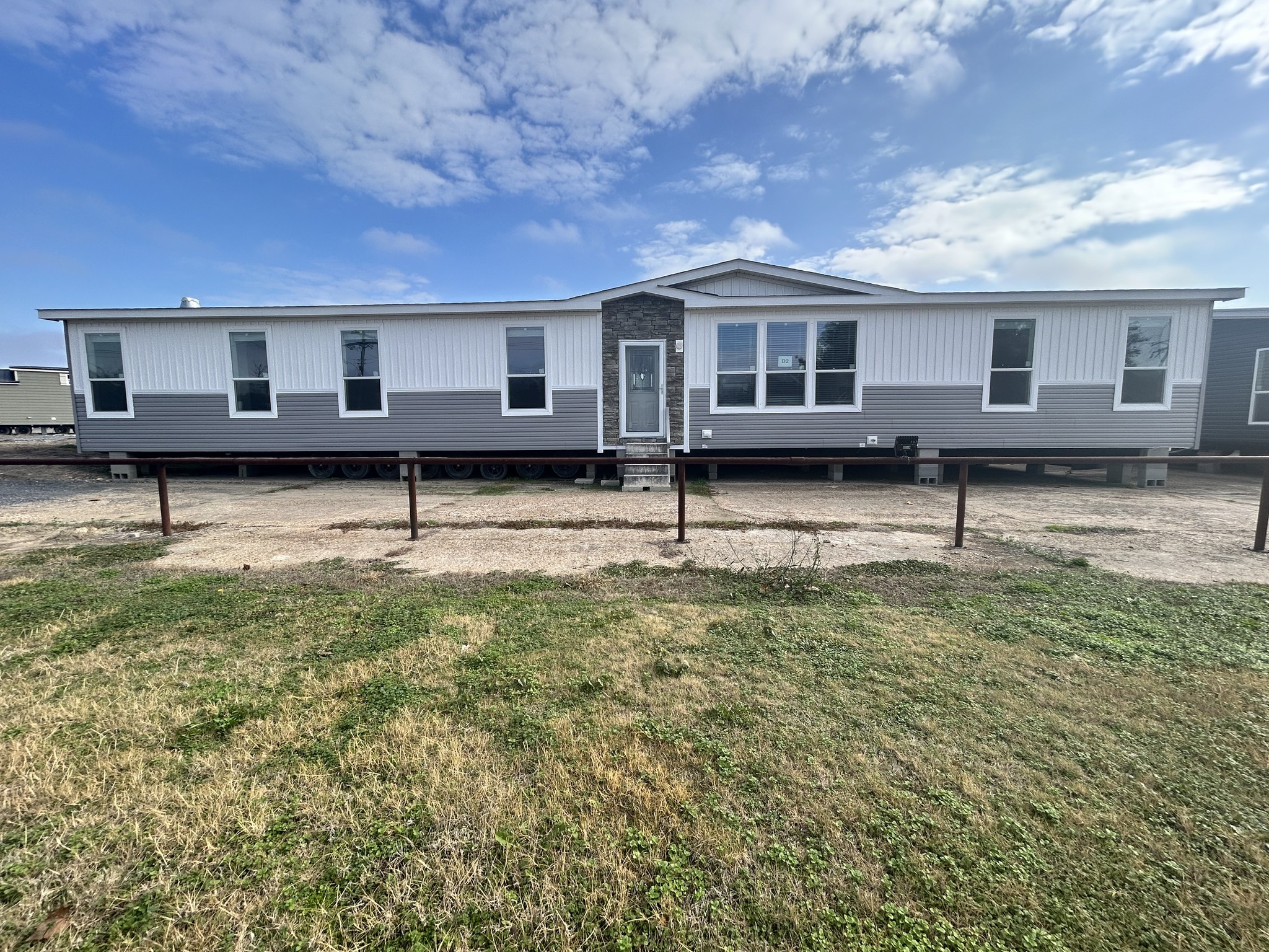 A single-story manufactured home with a gray and white exterior, featuring large windows and a central door. It sits on a gravel area with grass in the foreground under a partly cloudy blue sky.
