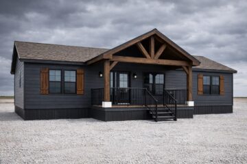 A modern, dark gray single-story house with wooden shutters and a large wooden porch under a cloudy sky. The setting is a gravel yard, evoking a rustic but contemporary feel.