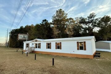 Single-story white mobile home with wooden accents, set on grassy terrain. Trees and power lines in the background under a blue sky with clouds.