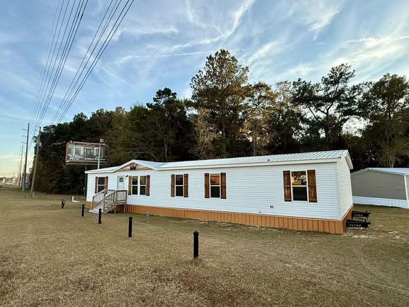 Single-story white mobile home with wooden accents, set on grassy terrain. Trees and power lines in the background under a blue sky with clouds.