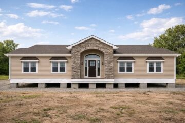 A tan, single-story house with a stone arch over the main entrance and dark brown door, surrounded by windows, stands on a gravel yard under a blue sky.