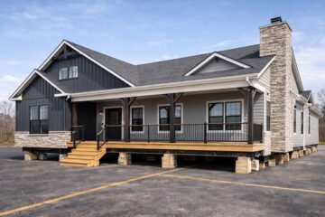 A modern modular house with dark siding and stone accents. It features a spacious porch with wooden steps and black railing, set on a sunny day.