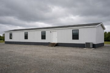 A white modular building with a gray foundation sits on a gravel lot under a cloudy sky. Three windows and a central door are visible, evoking a functional tone.