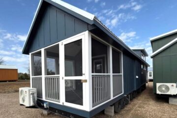 A compact, modern tiny house with dark blue siding and a white screened porch stands on gravel. The sky is clear and blue, conveying a serene atmosphere.