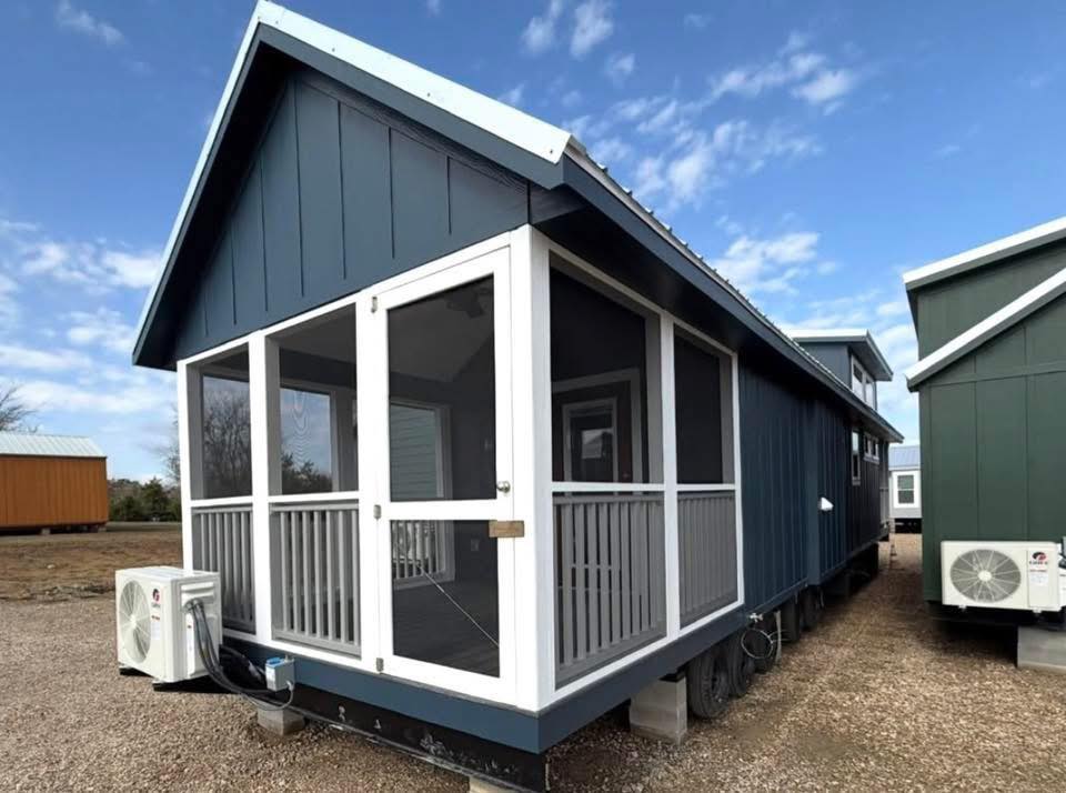 A compact, modern tiny house with dark blue siding and a white screened porch stands on gravel. The sky is clear and blue, conveying a serene atmosphere.