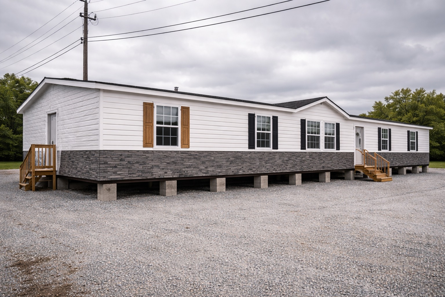 A white manufactured home with dark gray stone skirting and wooden steps sits on a gravel lot under an overcast sky, flanked by trees and power lines.