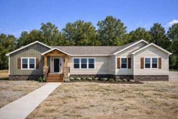 Single-story beige and green modular home with a gabled roof, wooden porch, and stone accents. Set against lush trees under a clear blue sky.