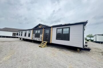 A large, beige manufactured home with black-framed windows and stone accents is set on gravel under a cloudy sky, conveying a sense of modernity.