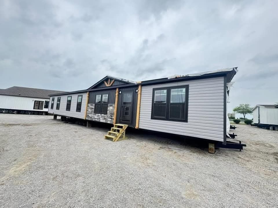 A large, beige manufactured home with black-framed windows and stone accents is set on gravel under a cloudy sky, conveying a sense of modernity.