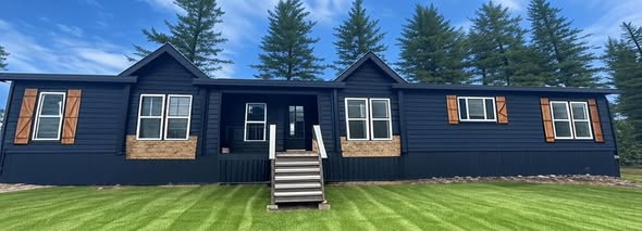 A modern, dark blue one-story house with wooden shutters and brick accents stands amidst lush green grass and tall pine trees under a clear blue sky.