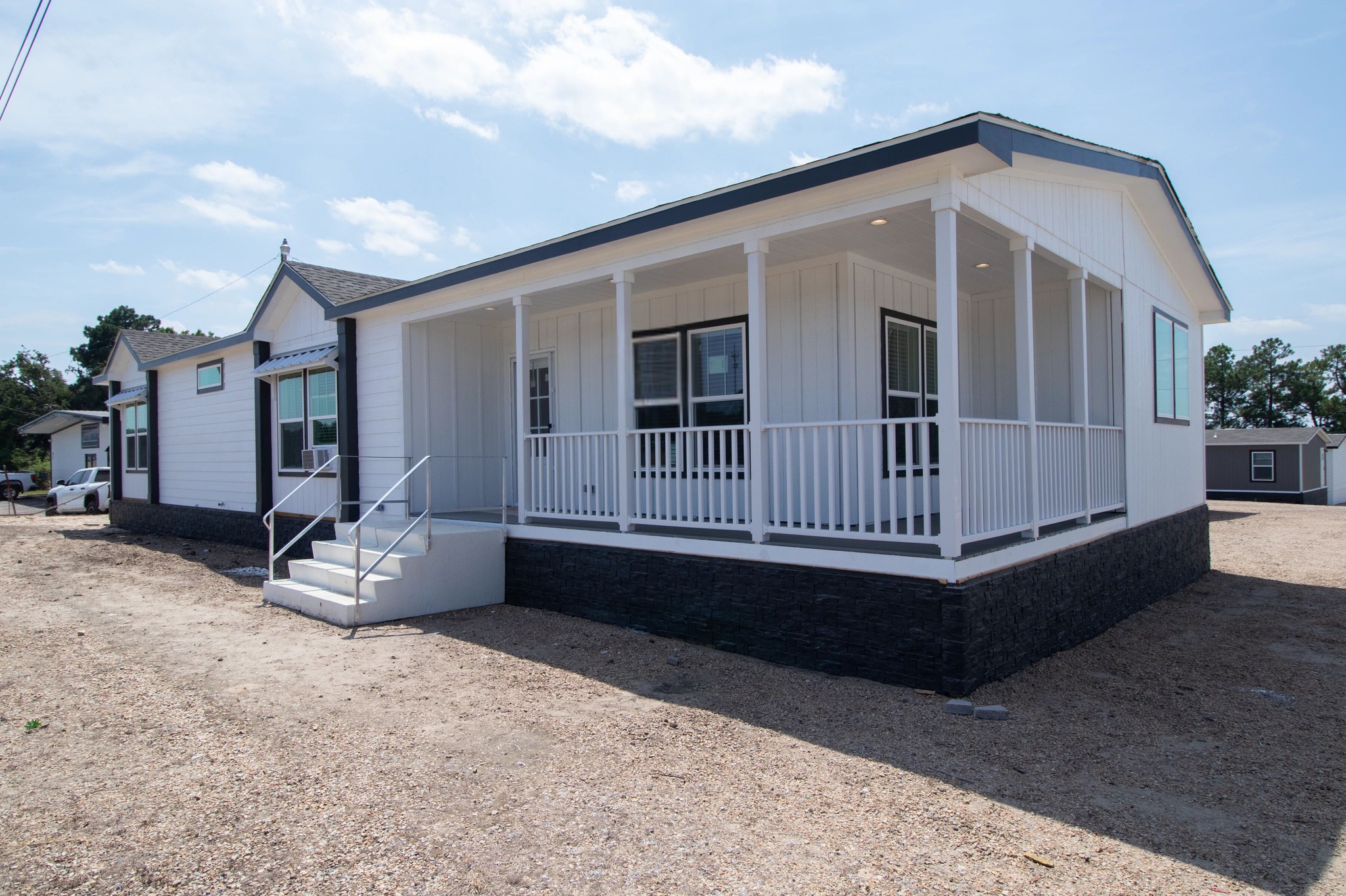 Modern white modular home with a covered porch and steps leading up, set on a gravel lot under a bright, partly cloudy sky; calm and inviting atmosphere.