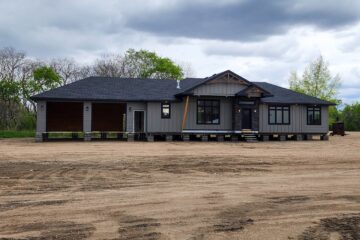 Single-story gray house with dark roofing on elevated supports, surrounded by sparse green trees under a cloudy sky. Two open garage bays on the left.