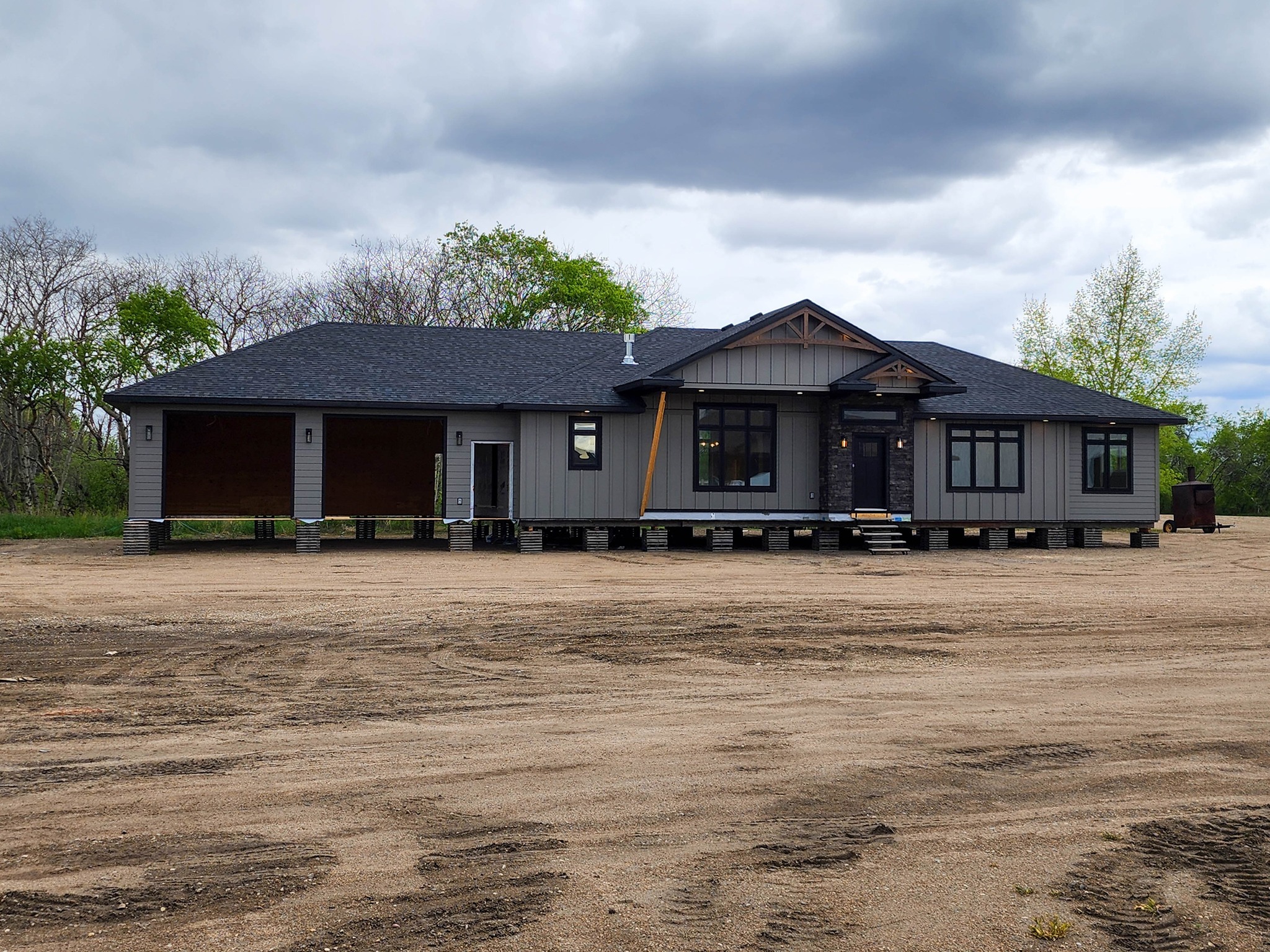 Single-story gray house with dark roofing on elevated supports, surrounded by sparse green trees under a cloudy sky. Two open garage bays on the left.