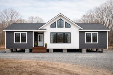 A modern, prefabricated gray and white house on a gravel lot with a gabled roof and large windows, surrounded by leafless trees under a cloudy sky.