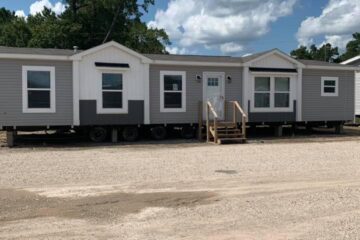 Modern gray and white mobile home on a gravel lot under a partly cloudy sky. The home has small wooden stairs and large windows, conveying a tranquil, open feel. Trees are visible in the background.