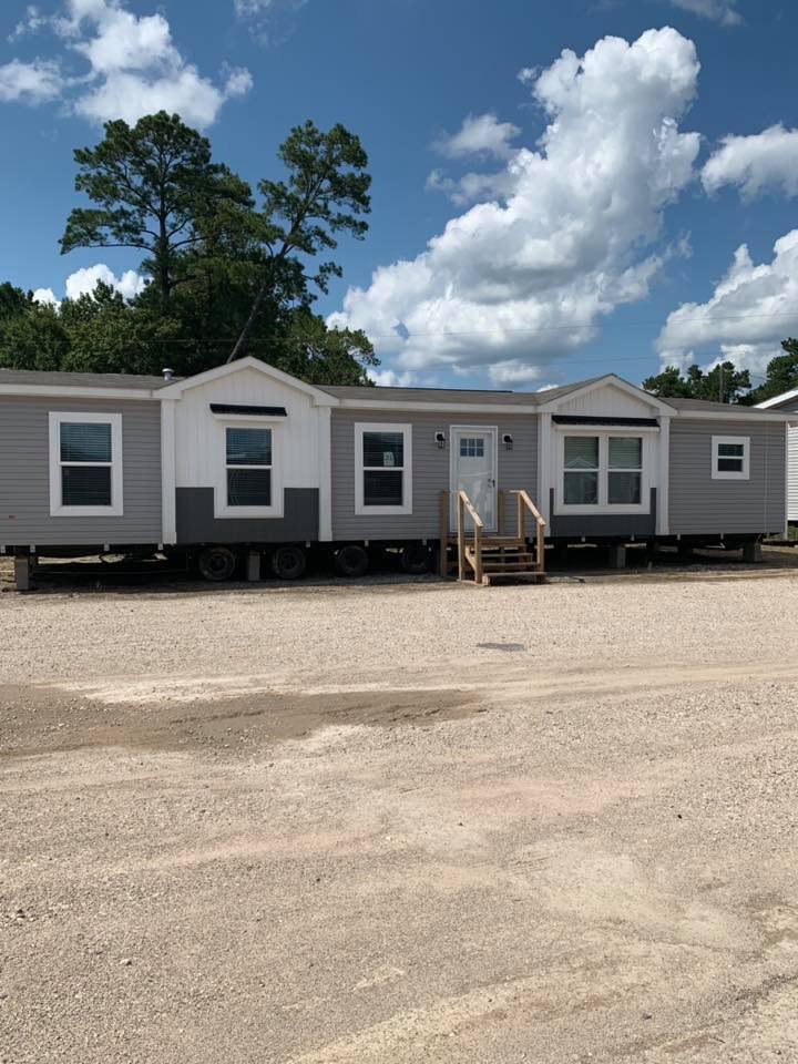 Modern gray and white mobile home on a gravel lot under a partly cloudy sky. The home has small wooden stairs and large windows, conveying a tranquil, open feel. Trees are visible in the background.