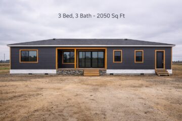 A modern, single-story modular home with gray siding and wooden trim, featuring large windows, a front porch, and two steps. Text above reads "3 Bed, 3 Bath - 2050 Sq Ft." The setting is overcast with an expansive, bare field.