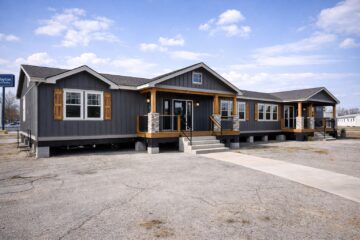 A modern, dark gray manufactured home with wooden accents and a welcoming front porch stands under a blue sky with scattered clouds.