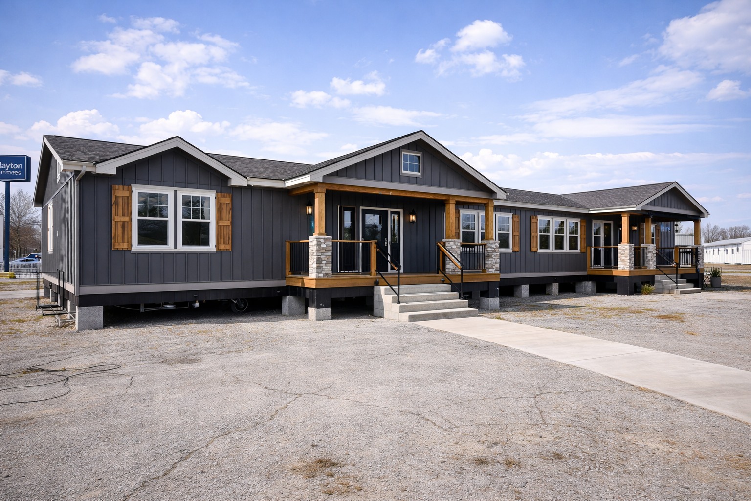 A modern, dark gray manufactured home with wooden accents and a welcoming front porch stands under a blue sky with scattered clouds.
