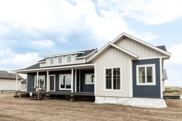 A modern, single-story house with blue and white siding, a large porch, and multiple windows, sits on a dirt lot under a partly cloudy sky.