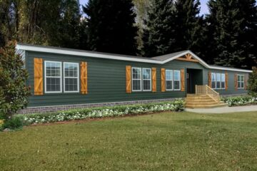 A long, green manufactured home with large windows and wooden shutters sits amidst a manicured lawn. Tall trees provide a serene backdrop.