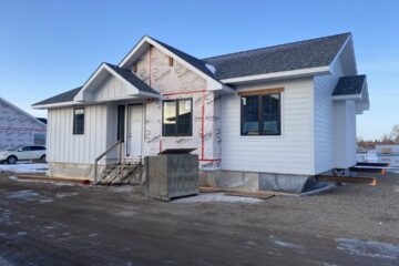 A partially constructed white house with unfinished siding and visible Tyvek wrap. A sign labeled "NO WOOD" is in front on a chilly, early morning.