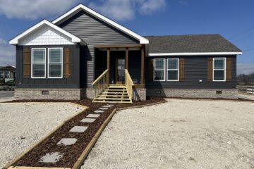 Single-story gray house with white trim and brown shutters, featuring a wooden porch. A stone path leads through a gravel yard under a bright, blue sky.