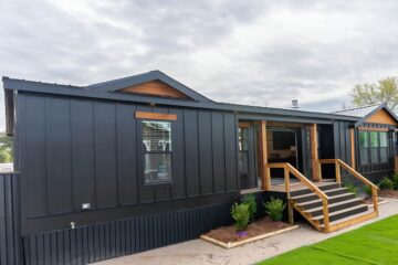 Stylish modern black modular home with wooden accents. Features a small porch with steps, situated under a cloudy sky, and surrounded by green plants.