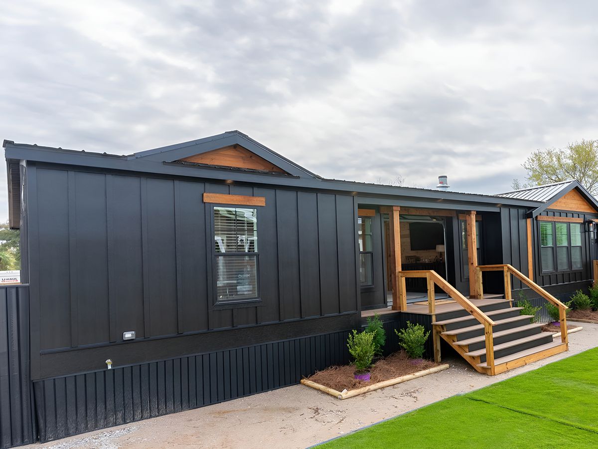 Stylish modern black modular home with wooden accents. Features a small porch with steps, situated under a cloudy sky, and surrounded by green plants.