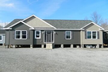 A single-story modular home on cinder blocks in a gravel lot. It features gray siding, white trim, and five windows, under a clear blue sky.