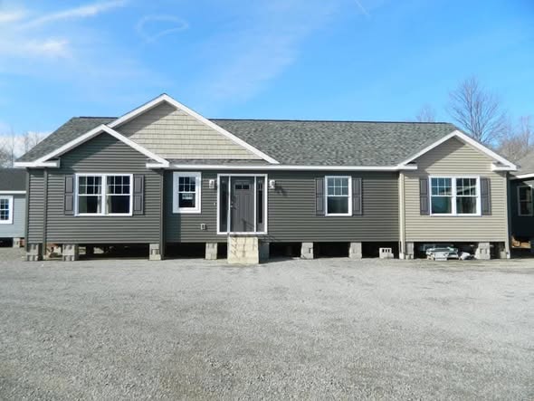 A single-story modular home on cinder blocks in a gravel lot. It features gray siding, white trim, and five windows, under a clear blue sky.