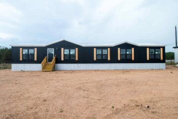A modern black mobile home with wooden shutters and a metal roof sits on a wide, sandy lot under a cloudy sky, creating a minimalist, serene atmosphere.
