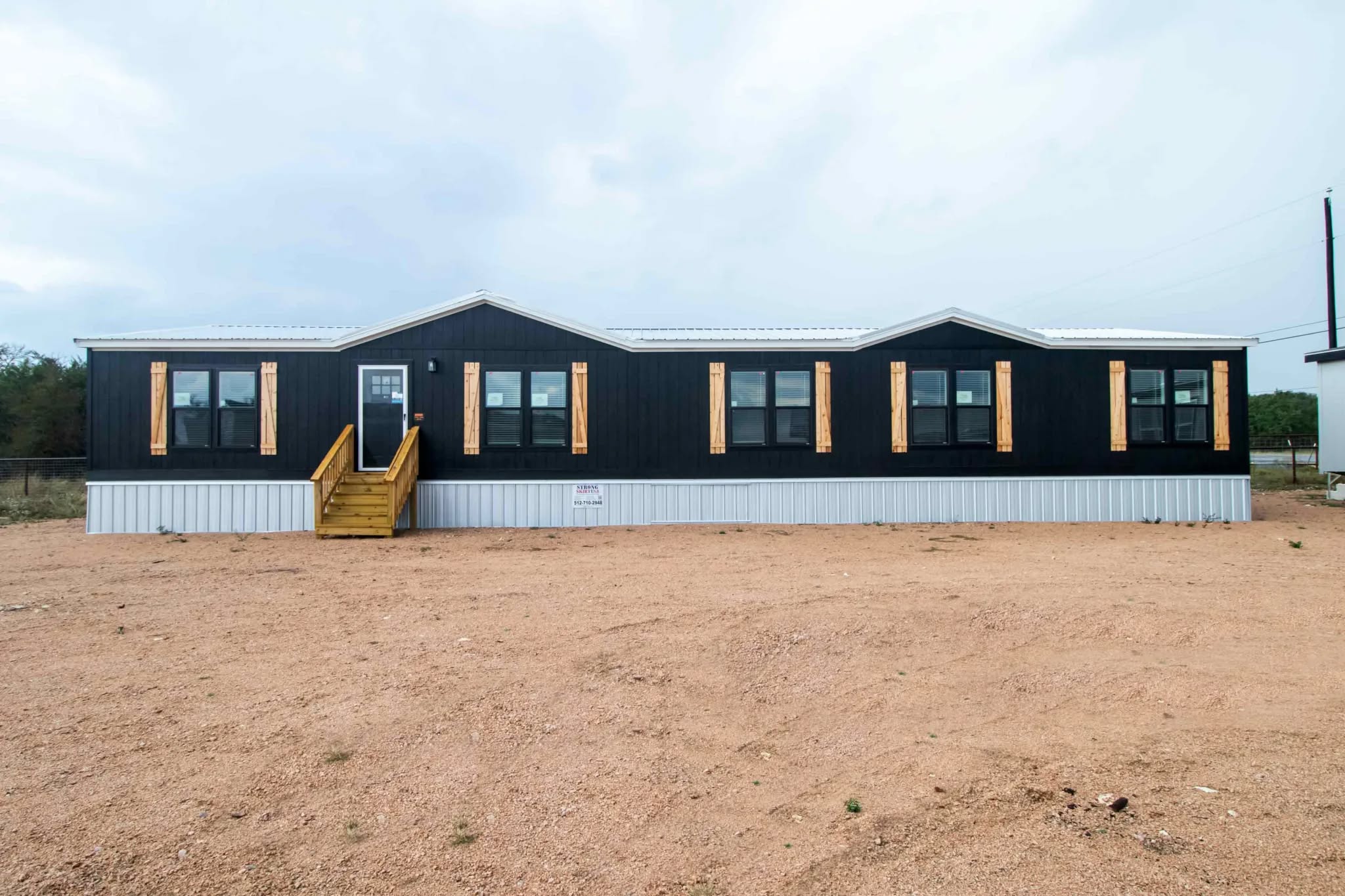 A modern black mobile home with wooden shutters and a metal roof sits on a wide, sandy lot under a cloudy sky, creating a minimalist, serene atmosphere.