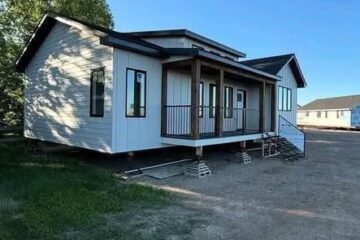 A small, white, single-story house with a pitched roof sits on a gravel area. It features a central porch and three steps, surrounded by grass and trees.