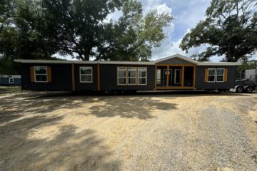 A modern manufactured home with dark siding and wooden trim sits on a sunny gravel lot, surrounded by tall trees and clear skies.