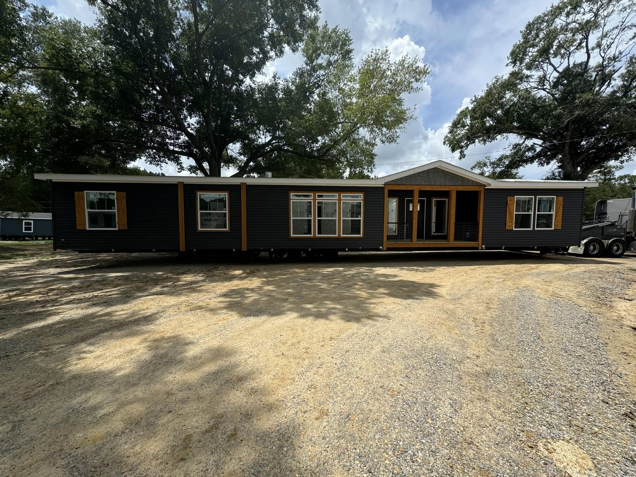 A modern manufactured home with dark siding and wooden trim sits on a sunny gravel lot, surrounded by tall trees and clear skies.