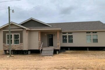 Single-story beige modular home on a dirt lot, with a covered front porch and stone accents. Overcast sky and a streetlamp are visible.