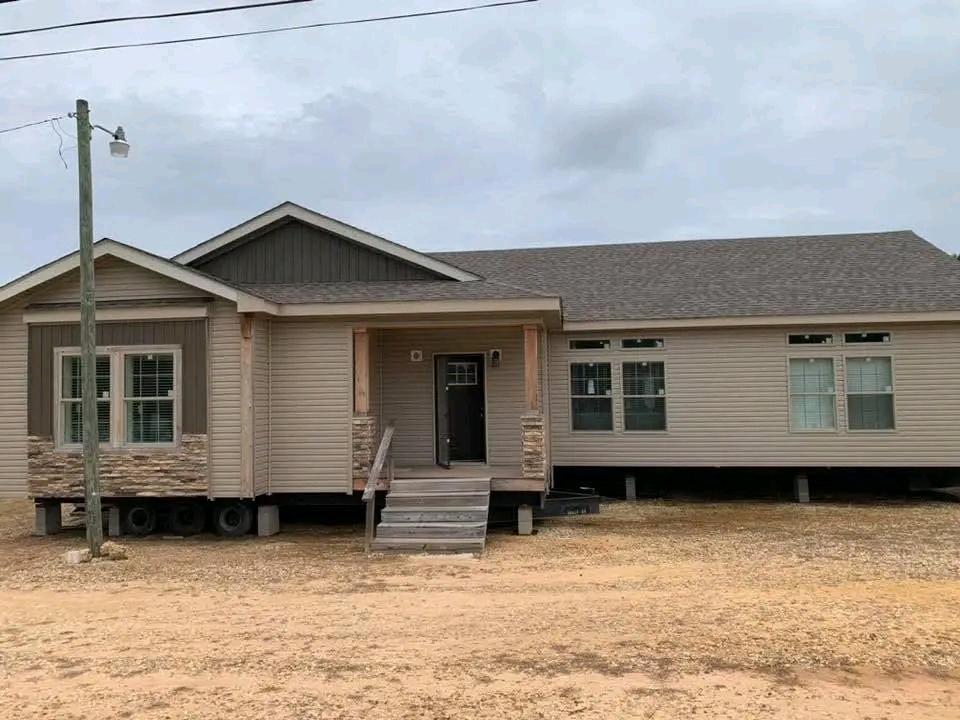 Single-story beige modular home on a dirt lot, with a covered front porch and stone accents. Overcast sky and a streetlamp are visible.