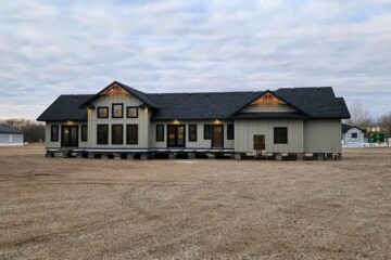 A single-story house with a black roof and light gray siding sits elevated on blocks in a gravel lot. Soft evening lights glow from the windows.
