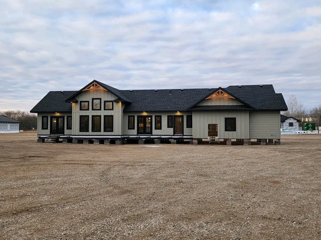 A single-story house with a black roof and light gray siding sits elevated on blocks in a gravel lot. Soft evening lights glow from the windows.