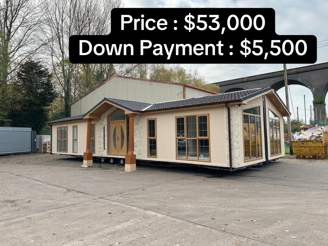 Mobile home for sale in a yard, priced at $53,000 with a $5,500 down payment. Surrounded by trees and industrial structures, overcast sky.