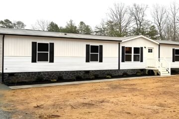 A white manufactured home with black shutters and a stone-patterned foundation. It sits on a sandy plot surrounded by leafless trees, conveying a serene rural setting.