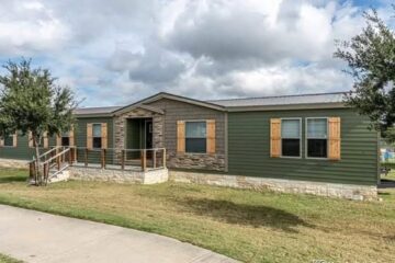 Single-story green manufactured home with stone accents, wooden shutters, a front ramp, and surrounded by grass and trees under a cloudy sky.