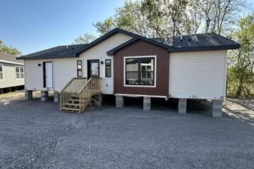 A newly constructed modular home on a gravel lot, elevated on concrete blocks. It features white siding, a brown accent panel, and a wooden staircase.