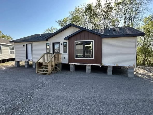 A newly constructed modular home on a gravel lot, elevated on concrete blocks. It features white siding, a brown accent panel, and a wooden staircase.