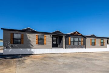 A modern manufactured home with gray siding and rustic wooden shutters sits under a clear blue sky. Its facade is accented with decorative stone.