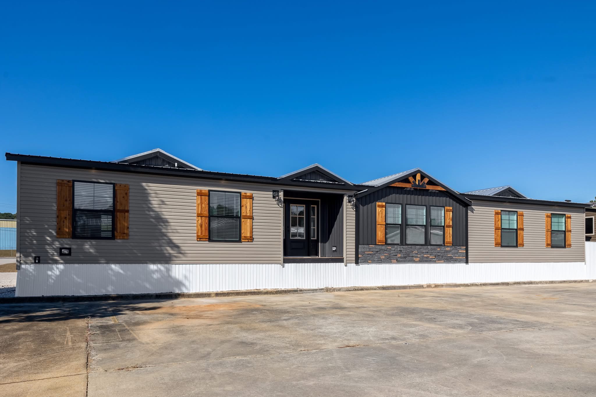 A modern manufactured home with gray siding and rustic wooden shutters sits under a clear blue sky. Its facade is accented with decorative stone.