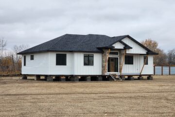 Single-story white house on blocks in a barren yard. Black roof, stone accent, and wooden scaffolding. Overcast sky, sparse trees, and a quiet, unfinished mood.